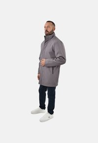 Giacca impermeabile grigia con colletto alto, maniche lunghe e tasche laterali con zip, abbinata a jeans di denim e sneakers bianche.