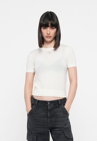 Weißer, kurzärmeliger, cropped Pullover mit tailliertem Schnitt, rundem Halsausschnitt und kleinem Logodetail. Kombiniert mit dunkelgrauen Cargo-Shorts.