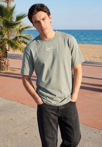 Mann mit dunklen Haaren, der ein hellgraues T-Shirt und schwarze Hose trägt, steht mit den Händen in den Taschen an der Strandpromenade.