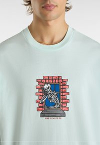 Maglietta azzurra a maniche corte con una grafica di uno scheletro in una finestra di un muro di mattoni, con il testo "MORIRE PER PARLARTI" sotto.