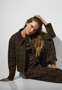 Femme aux longs cheveux blonds assise par terre, portant une veste à carreaux marron foncé et un pantalon assorti, reposant une main sur sa tête.