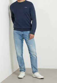 Mann trägt einen marineblauen Guess-Pullover, hellblaue Jeans mit leichten Abnutzungserscheinungen und weiße Sneaker, während er gegen eine weiße Wand steht.