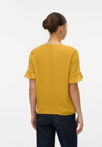 Blusa gialla senape a maniche corte con orlo arricciato, scollatura rotonda, realizzata in tessuto morbido, abbinata a jeans blu scuro. Vista posteriore.
