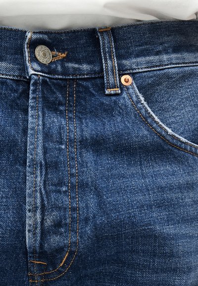 Κοντινό πλάνο μπροστινού μέρους μπλε τζιν με ορατό κουμπί, θηλιά για ζώνη, μπροστινή τσέπη και πορτοκαλί ραφές.