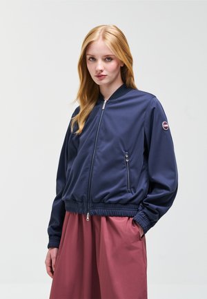 Jeune femme aux longs cheveux blonds portant une veste aviateur bleu marine zippée et une jupe taille haute rose poussiére, debout devant un fond uni.