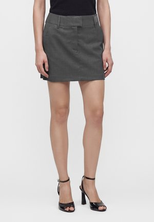 Femme portant une mini-jupe gris foncé avec passants pour ceinture et un haut noir sans manches, associée à des sandales noires à talons hauts et orteils découverts.