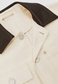 Cremefarvet skjorte med en brun læderkrave, to knaplukninger og en brystlomme. Struktureret stof med subtile spidser.