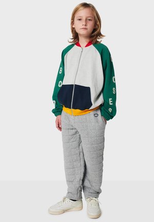 Barn står klädd i en färgblockerande zip-up jacka med gröna ärmar och grå vadderade byxor samt vita sneakers framför en enkel bakgrund.