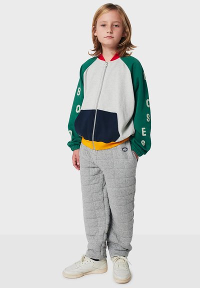 Child standing wearing a color-block zip-up jacket with green sleeves and gray quilted pants and white sneakers on a plain background.