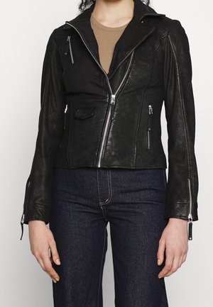 Mujer con chaqueta biker de cuero negra ajustada con bolsillos con cremallera y vaqueros azul oscuro de tiro alto, de pie frente a un fondo simple.