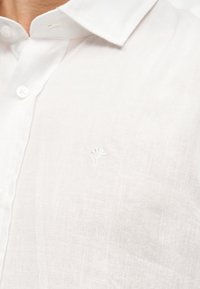 Gros plan d'un col de chemise blanche à boutons et de la partie supérieure de la poitrine avec un petit logo brodé discret sur le côté gauche.