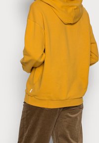 Sudadera con capucha de color mostaza en tela suave, con un ajuste relajado y puños acanalados, combinada con pantalones de pana marrones.