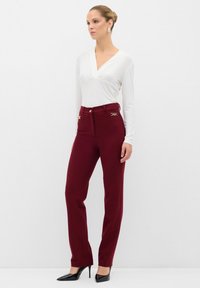 Witte lange mouwen top met rouleren aan de zijkant; bordeauxrode high-waisted broek met gouden accenten op de zakken; model draagt zwarte puntige hakken.