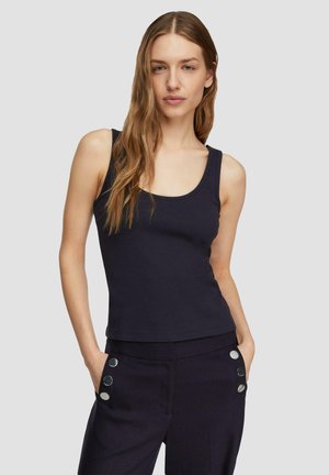 Młoda kobieta o długich brązowych włosach, ubrana w czarny top bez rękawów i czarne spodnie z srebrnymi guzikami, z rękami w kieszeniach.