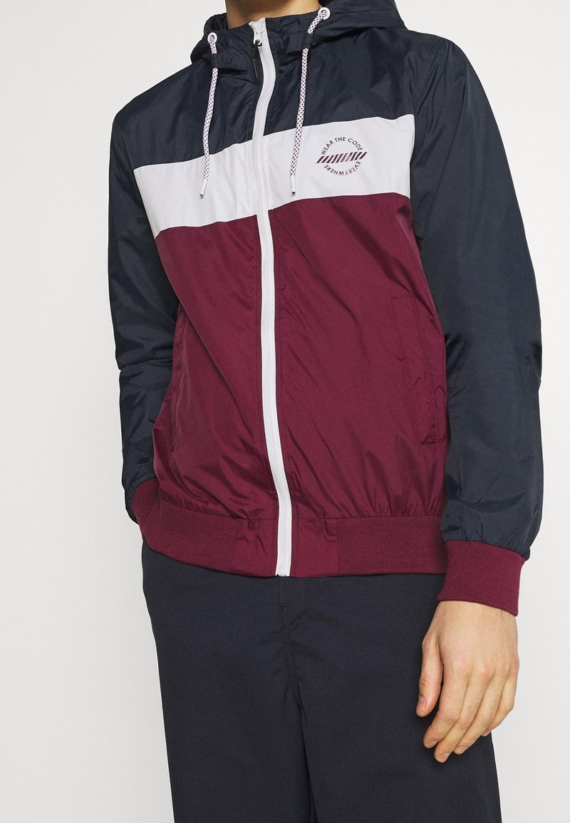 Homme portant une veste coupe-vent bicolore avec des parties bleu marine, blanc et bordeaux, fermeture éclair à l'avant, capuche et cordons, debout les mains basses.