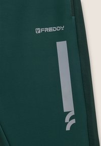 Pantalons de sport verts avec un panneau latéral texturé, un logo Freddy argenté bien visible et une bande verticale argentée.