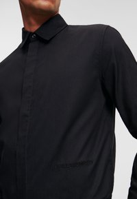 Schwarzes Langarmshirt mit strukturiertem Kragen, glatter Textur, dezenter gemusterter Stoff und gesticktem "KARL LAGERFELD"-Detail am Saum.