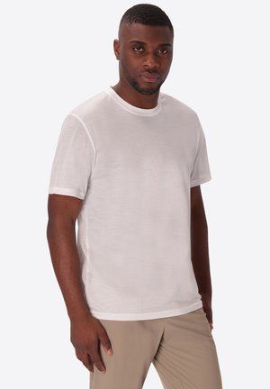 Uomo che indossa una maglietta bianca semplice a maniche corte e pantaloni beige, in piedi contro uno sfondo grigio chiaro, che guarda avanti.