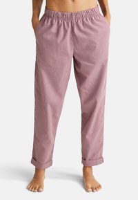 Persoon in rood-witte geruite losse broek met elastische tailleband, handen in de zakken, en blote voeten op een witte achtergrond.