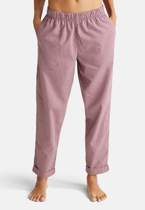 Persoon in rood-witte geruite losse broek met elastische tailleband, handen in de zakken, en blote voeten op een witte achtergrond.