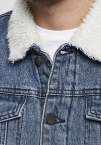 Hombre vistiendo una chaqueta de mezclilla azul con cuello de sherpa blanco sobre una camiseta blanca de cuello redondo, mostrando la parte superior del pecho y la zona del cuello.