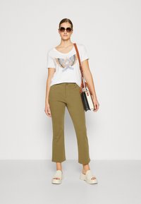 Bílá tričko s grafikou orla, olivově zelené zkrácené kalhoty a světle béžové platformové sandály. Hnědá crossbody taška s černobílými akcenty.