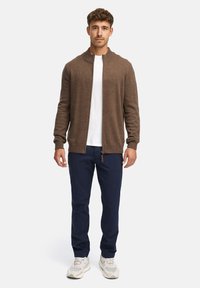Cardigan zippé marron en matériau doux, avec un col et des poignets côtelés. Porté par-dessus un T-shirt blanc et un pantalon bleu marine.