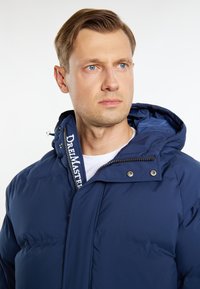 Marineblå pufferjakke med hætte, lynlås og logo på kraven. Har en glat overflade og polstret design for varme.
