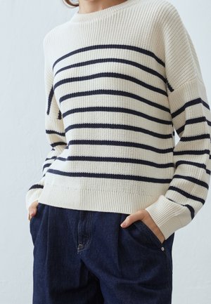 Personne portant un pull en tricot crème à rayures horizontales noires et un pantalon en denim bleu foncé, mains dans les poches, sur un fond clair uni.