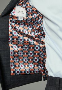 Sakó na mieru s tmavým textúrovaným povrchom. Podšité vzorovanou látkou so geometrickými tvarmi v oranžovej, námorníckej modrej a bordovej farbe.
