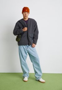 Grå fleecejakke med to forlommer, gult logo på en orange hue, lyseblå løse jeans og hvide sneakers med gummisåler.