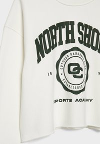 Sweat-shirt blanc à manches longues avec texte noir "NORTH SHORE" et un logo circulaire indiquant "Outdoor Handball Club Established 1985 Sports Academy."