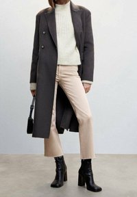 Longue manteau en laine gris sur un pull léger, associé à un jean beige cut-off et des bottes noires à talons. Petit sac à main noir en accent.