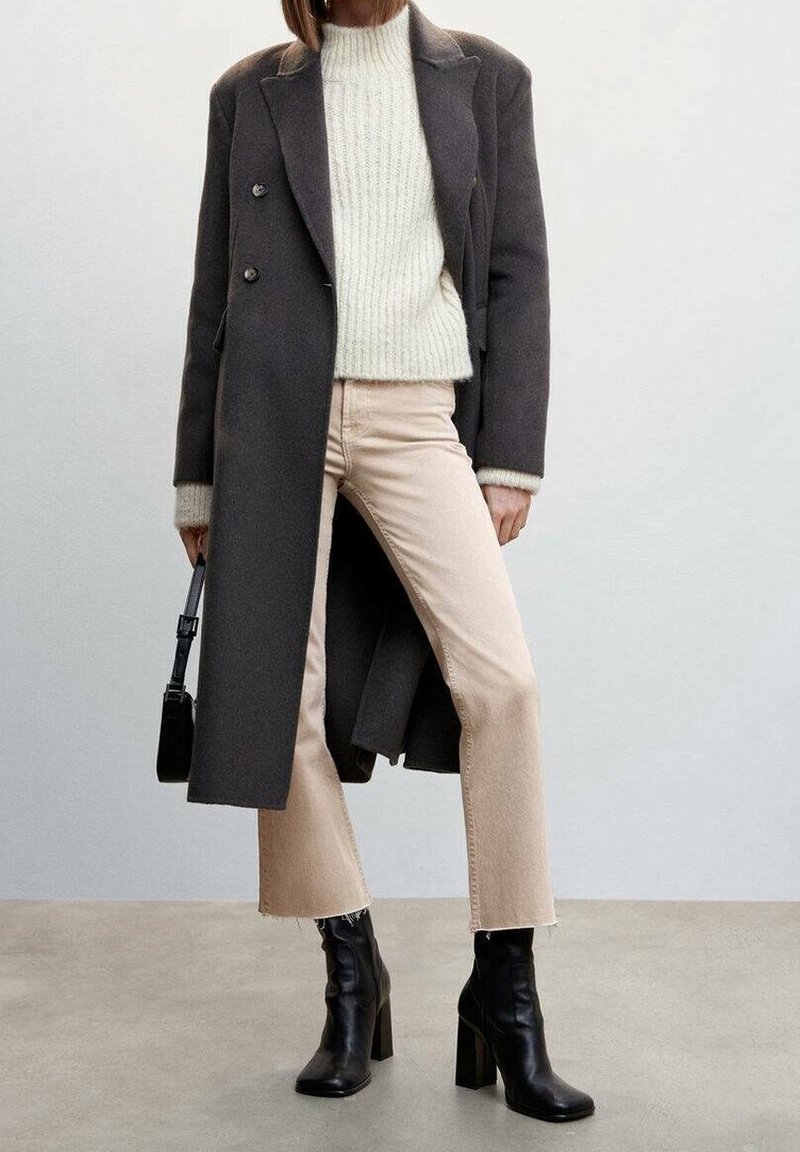 Longue manteau en laine gris sur un pull léger, associé à un jean beige cut-off et des bottes noires à talons. Petit sac à main noir en accent.