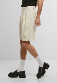 Shorts plissés couleur crème avec une taille élastique, dotés de poches latérales, portés avec des chaussettes blanches et des mocassins noirs épais.