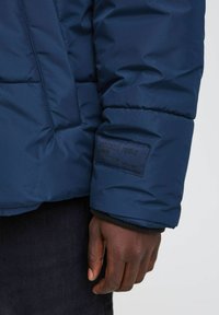 Marinblå pufferjacka med en strukturerad yta, med en patch på ärmen som säger "DEDICATED TO DENIM PERFECTION." Svart ribbad manschett.