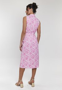 Abito floreale rosa con silhouette aderente, collo alto e design senza maniche. Il motivo presenta grandi fiori e foglie. Sandali con tacco oro.