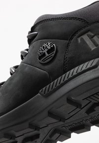 Schwarze Wildleder-Sneaker mit strukturierten Akzenten, ausgestattet mit einem markanten Logo und einer robusten Gummisohle. Schnürsenkel, die mit schwarzen Ösen gesichert sind.