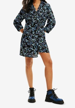 Woman wearing a black floral wrap dress with blue and white flowers and blue and black lace-up ankle boots, standing with hands on hips.