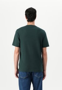 Zöld pamut t-shirt, kerek nyakkivágással és rövid ujjakkal, hátulról látható, kék farmernadrággal párosítva. Sima textúra.