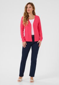 Roze cardigan, open voorkant, lange mouwen, gedragen over een witte top, gecombineerd met donkerblauwe jeans en beige hakken. Soepele textuur, casual stijl.