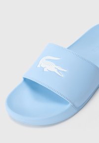 Sabo de plajă de culoare albastră deschisă cu logo-ul alb de crocodil pe banda lată și tălpica texturată pe fundal simplu.