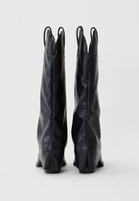 Paire de bottes mi-mollet en cuir noir avec languettes de traction, debout contre un fond gris clair uni.