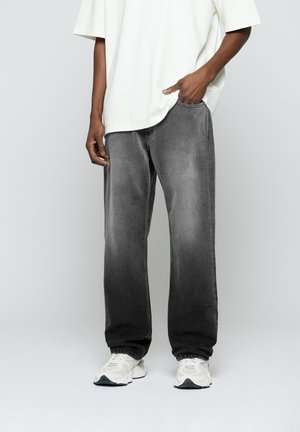 Personne portant un jean gris ample, un t-shirt blanc oversize et des baskets blanches, debout avec une main dans la poche devant un fond uni.