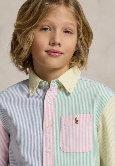 Παιδί με μεσαίου μήκους ξανθά μαλλιά που φοράει πολύχρωμο ριγέ πουκάμισο με κουμπιά και τσεπάκι στο στήθος με λογότυπο αλόγου.
