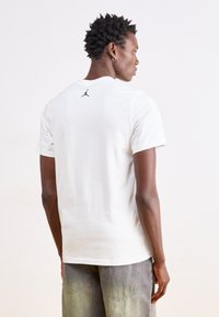 Λευκή βαμβακερή μπλούζα με στρογγυλή λαιμόκοψη, κοντά μανίκια και ένα μικρό μαύρο λογότυπο Jordan στο πίσω μέρος του λαιμού. Το ύφασμα φαίνεται απαλό και λείο.