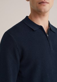 Tamsiai mėlynas megztas polo marškinėlis su tekstūruota paviršiaus struktūra, trumpu apykakle ir pusiau užtrauktuku priekyje. Akcentuotas prigludęs siluetas.