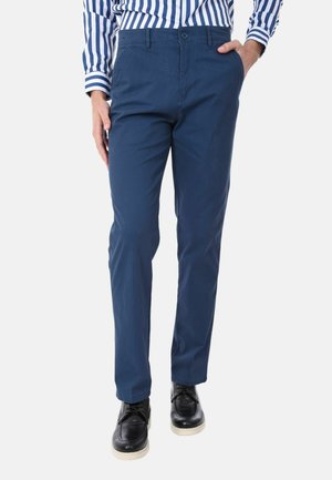 Uomo che indossa pantaloni chino blu, una camicia a righe blu e bianche e scarpe eleganti nere, in piedi con una mano in tasca.