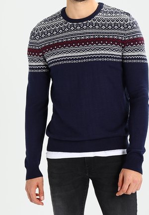 Man draagt een marineblauwe trui met witte en bordeauxrode gestreepte patronen over de borst en bovenarmen, gecombineerd met een zwarte broek.