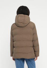 Puffjakke i brunt stof, med hætte og quiltet design. Bagudsigten viser en afslappet pasform og elastiske manchetter.